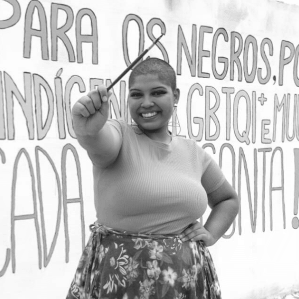 Foto em preto e branco de Beatriz, jovem negra de cabelo raspado. Ela veste uma camiseta e uma saia florida e segura um pincel. Atrás dela está o painel que a jovem pintou