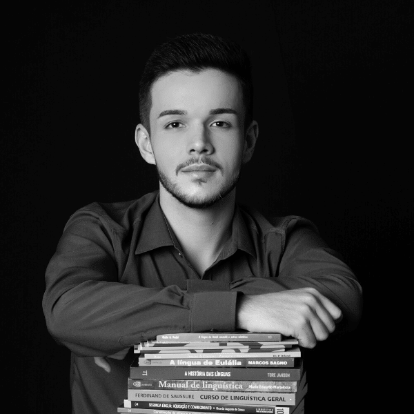 Foto em preto e branco de Felipe, jovem branco com barba e bigode. Ele está apoiado em uma pilha de livros