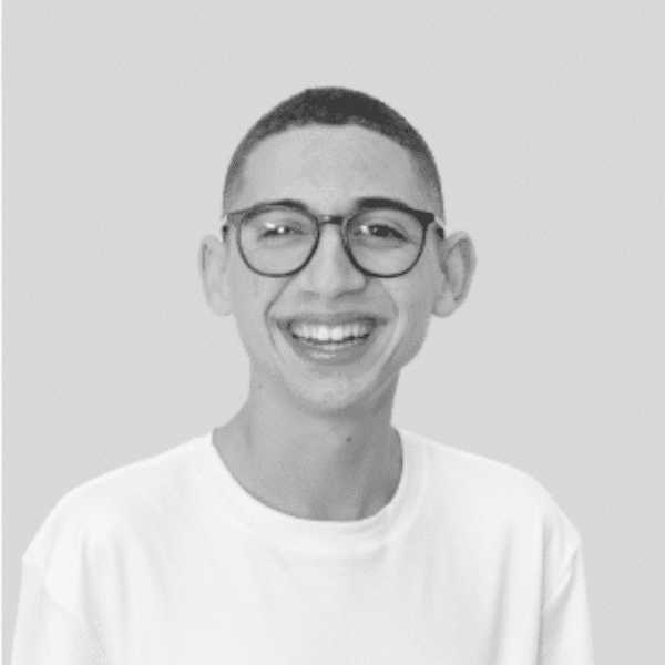 Retrato em preto e branco de um menino sorrindo para a câmera. Ele tem cabelo curto, usa óculos de armação redonda e uma camiseta branca.
