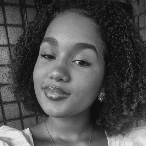 Retrato em preto e branco de uma menina sorrindo. Ela tem cabelos curtos e cacheados, usa brincos em formato de flor e uma corrente delicada.