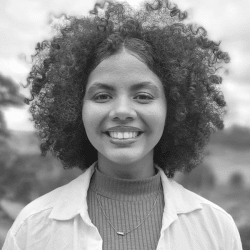 Foto em preto e branco de Débora, uma jovem negra de cabelos crespos. Ela está ao ar livre e sorri para a foto. Ela veste uma blusa de gola alta com uma camisa social aberta