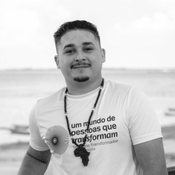 Foto em preto e branco de Jeferson, jovem de cabelo curto que sorri para a foto. Ele tem barba, bigode e usa uma camiseta em que se lê "um mundo de pessoas que transformam". Ele também usa um colar com o formato do continente africano e está ao ar livre, onde se vê um rio