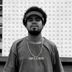 Foto em preto e branco de Jonathas, jovem negro de cabelos curtos e crespos com barba e bigode. Ele usa uma viseira, camiseta e corrente