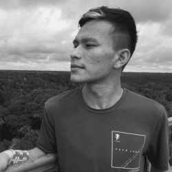 Foto em preto e branco de Luiz Henrique, jovem indígena vestindo uma camiseta e olhando para o horizonte. Ao fundo está o céu e a Floresta Amazônica