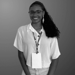 Foto em preto e branco de uma jovem de cabelo preso, cacheado e longo, está sorrindo para foto e usando uma camiseta branca, óculos de grau e um crachá