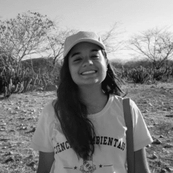 A imagem em preto e branco mostra Lara sorridente, usando boné e vestindo uma camiseta com as palavras "Ciências Ambientais" estampadas. Ela está ao ar livre, em uma área de vegetação seca com árvores e cactos ao fundo, característicos de uma região de clima árido ou semiárido.