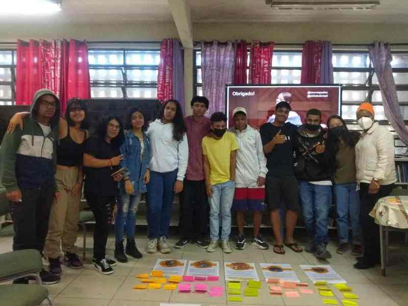 Jovens enfileirados posando para foto. No chão à frente do grupo, há folhas de papel e notas adesivas