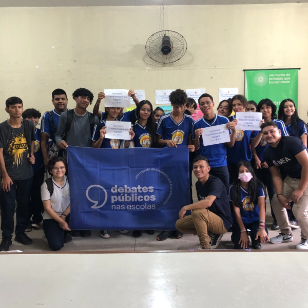 Grupo de jovens em uma sala de aula sorriem para a foto em volta da bandeira dos Debates Públicos nas Escolas