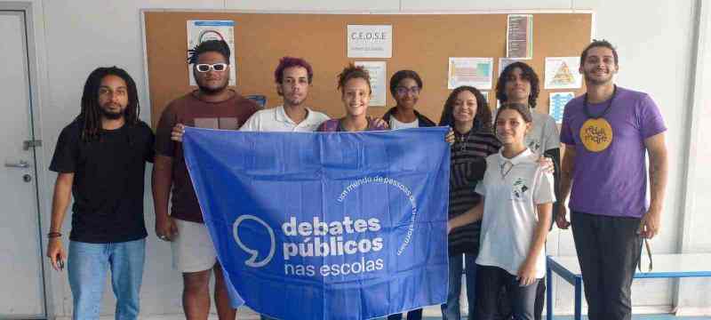 Pequeno grupo de pessoas reunidas em uma sala seguram a bandeira dos Debates Públicos nas Escolas