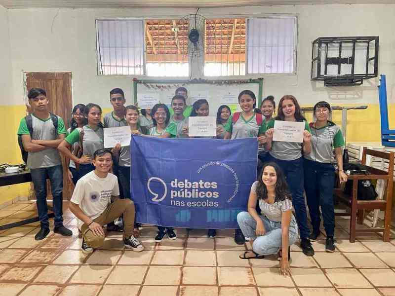 Grupo de jovens reunidos em um pátio. A turma sorri para a foto e mostra a bandeira dos Debates Públicos nas Escolas