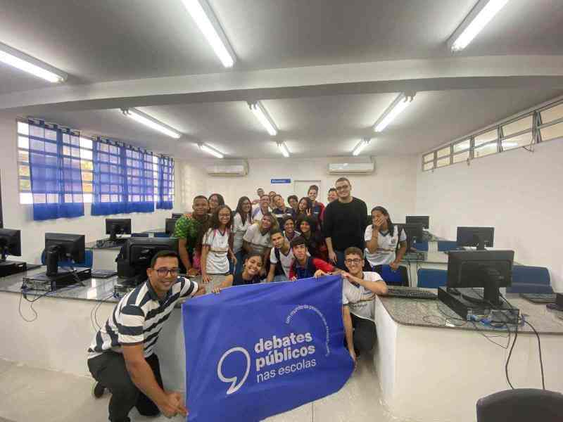 Grupo reunido em um laboratório de informática sorri para a foto. As pessoas à frente seguram uma bandeira azul com os dizeres "Debates Públicos nas Escolas"