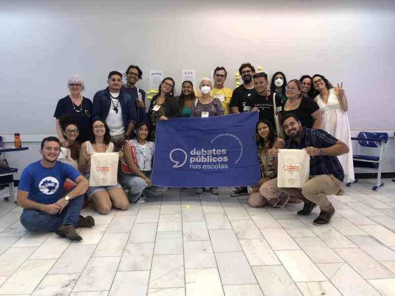 Grupo de pessoas reunidas sorrindo para a foto em uma sala de aula. Ao centro, algumas delas seguram uma bandeira azul escuro escrito "Debates Públicos nas Escolas"