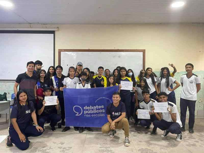 Grupo de jovens em uma sala de aula sorriem para a foto em volta da bandeira dos Debates Públicos nas Escolas