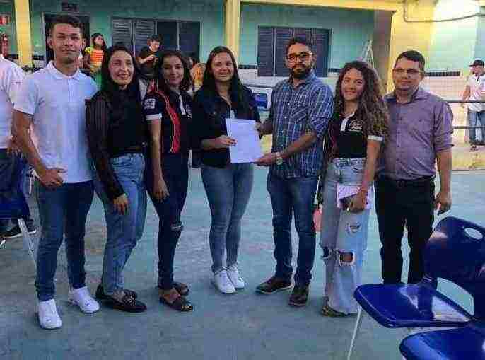 Um grupo de jovens junto a um adulto posam para a foto em um pátio de escola. A jovem no centro entrega um papel para o adulto