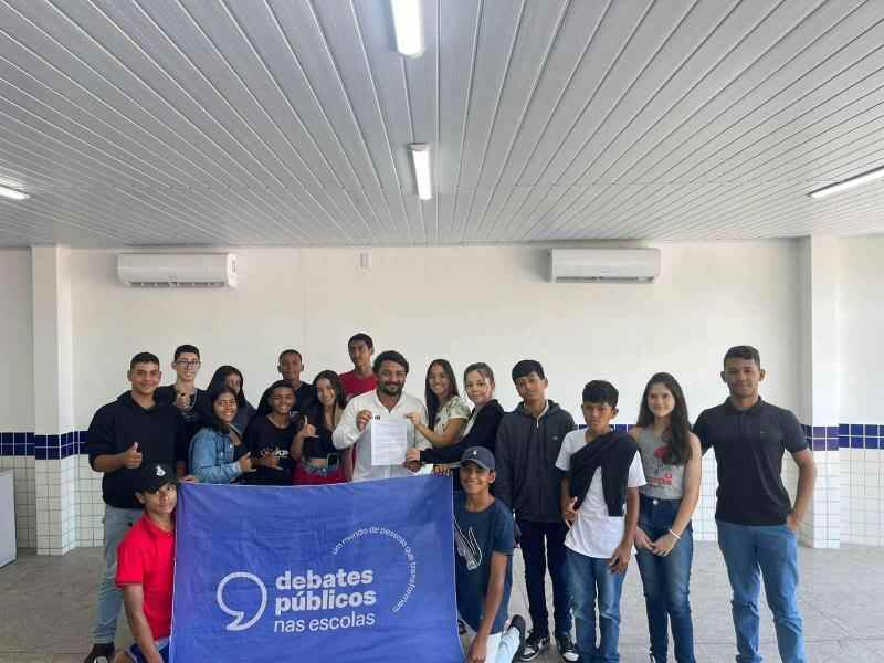 Grupo de jovens reunidos em uma sala de aula. A turma sorri para a foto e mostra a bandeira dos Debates Públicos nas Escolas