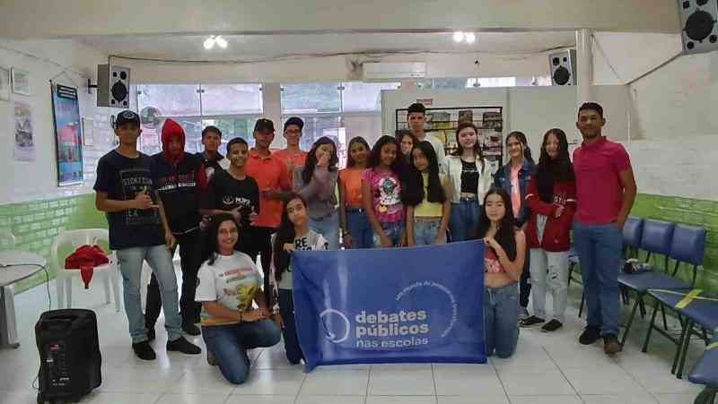 Grupo de jovens reunidos em uma sala de aula. A turma sorri para a foto e mostra a bandeira dos Debates Públicos nas Escolas