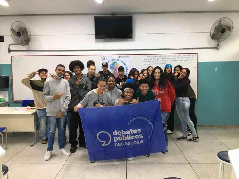 Grupo de jovens reunidos em uma sala de aula. A turma sorri para a foto e mostra a bandeira dos Debates Públicos nas Escolas