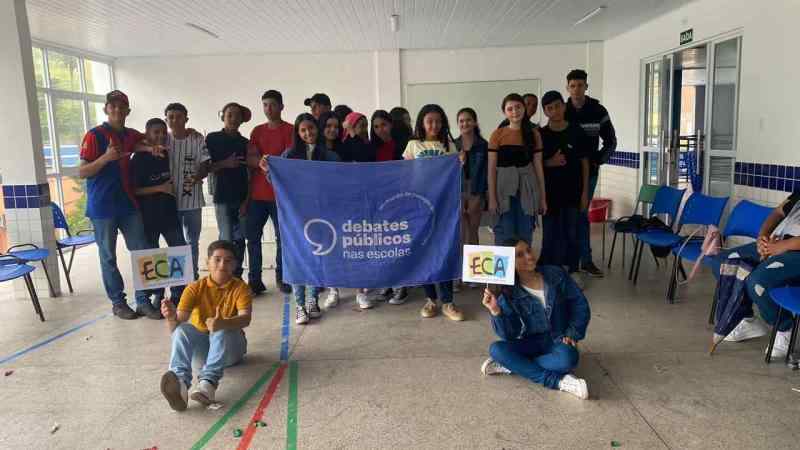 Grupo de jovens reunidos em uma sala de aula. A turma posa para a foto e segura a bandeira dos Debates Públicos nas Escolas. Dois estudantes estão sentados à frente segurando placas em que se lê "ECA"