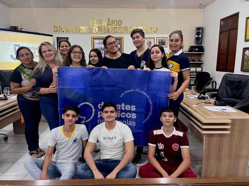 Grupo de pessoas reunidas em um plenário. Elas sorriem para a foto e seguram a bandeira dos Debates Públicos nas Escolas