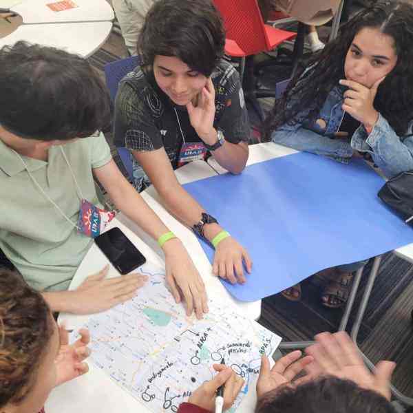 Jovens reunidos em torno de uma mesa. Sobre a mesa, há um cartaz e um mapa. O grupo está observando o mapa e fazendo anotações