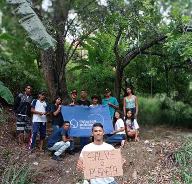 Grupo de jovens reunidos em uma área de floresta. Ao fundo, um grupo posa para a foto e segura uma bandeira azul dos Debates Públicos nas Escolas. À frente, um jovem segura uma placa de papelão com os dizeres "SALVE O PLANETA"