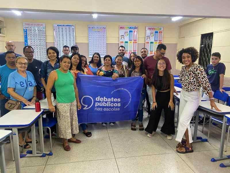 Grupo de pessoas reunidas em uma sala de aula. As pessoas sorriem para a foto e seguram uma bandeira azul dos Debates Públicos nas Escolas