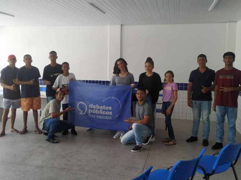 Grupo de jovens em uma sala de aula sorriem para a foto em volta da bandeira dos Debates Públicos nas Escolas