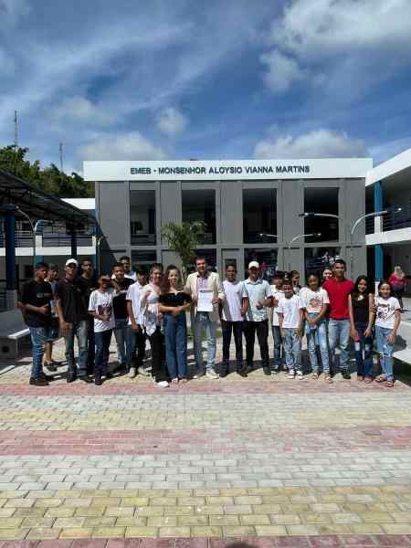 Grupo de jovens reunidos em um pátio. Ao centro, há um adulto mostrando uma folha de papel. Ao fundo, há um prédio baixo na cor cinza onde se lê "EMEB - Monsenhor Aloysio Vianna Martins"