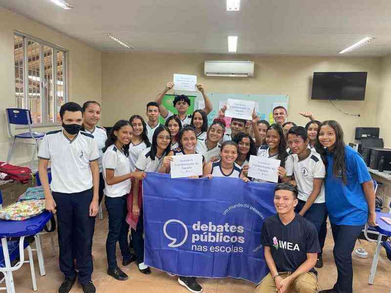 Grupo de jovens reunidos em uma sala de aula. A turma sorri para a foto e mostra a bandeira dos Debates Públicos nas Escolas