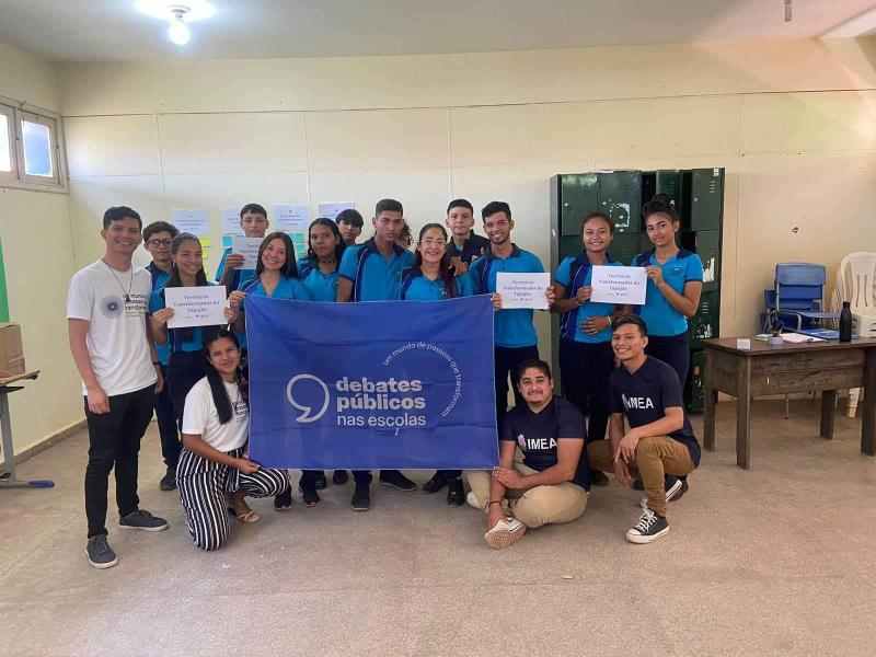 Grupo de jovens em uma sala de aula sorriem para a foto em volta da bandeira dos Debates Públicos nas Escolas