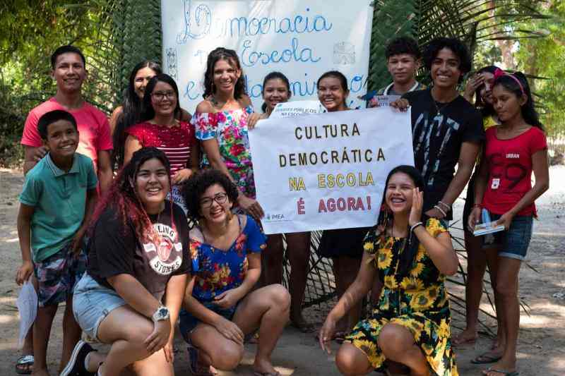 Grupo de jovens reunidos ao ar livre seguram cartazes sobre cultura democrática