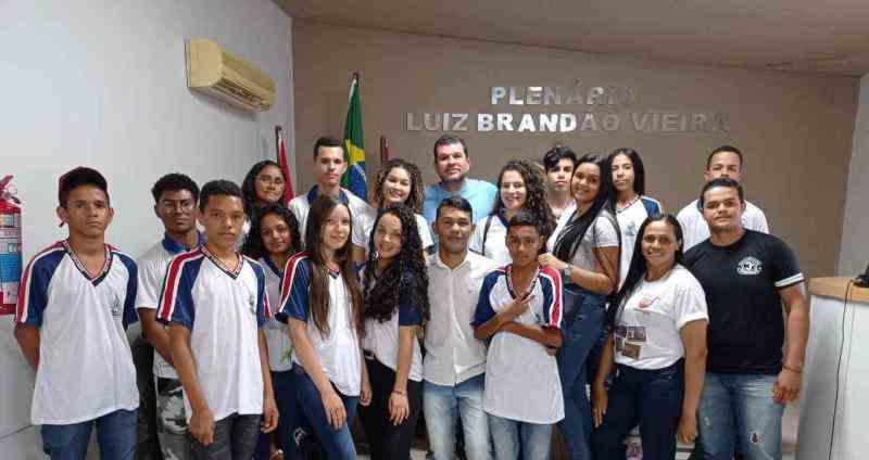 Grupo de jovens posando para a foto, a maioria veste uniformes escolares. No centro do grupo, há um homem adulto vestindo camiseta azul clara e um jovem com camisa social. O grupo está em uma sala, onde ao fundo é possível ver uma bandeira do Brasil e os dizeres "Plenário Luiz Brandão Vieira", em uma parede bege