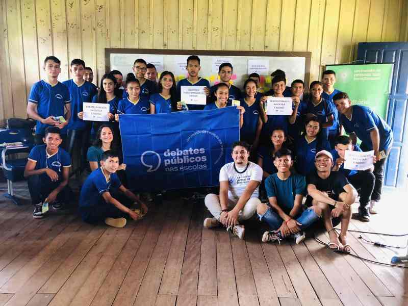 Grupo de jovens em uma sala de aula sorriem para a foto em volta da bandeira dos Debates Públicos nas Escolas