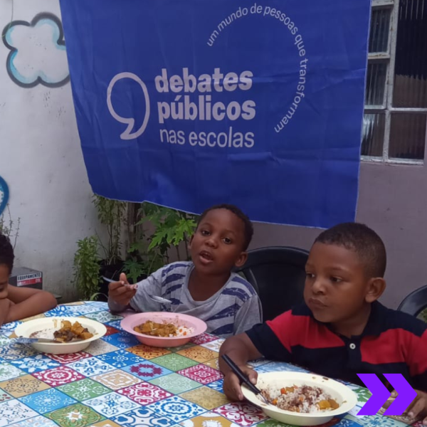 há duas crianças sentadas em uma mesa com seus respectivos pratos de comida, se alimentando. Logo atrás deles, há uma bandeira roxa dos Debates Públicos nas Escolas. 