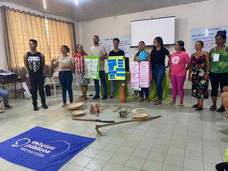  diversas pessoas segurando cartazes com ferramentas para mexer com a terra espalhadas no chão à sua frente e logo em seguida a bandeira dos Debates Públicos nas Escolas