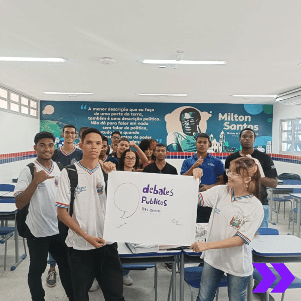 A imagem mostra um grupo de estudantes em uma sala de aula segurando um cartaz que diz "Debates Públicos nas Escolas". Ao fundo, há uma parede com uma ilustração de Milton Santos e uma citação dele sobre política e educação. Os alunos estão sorrindo e posam para a foto.