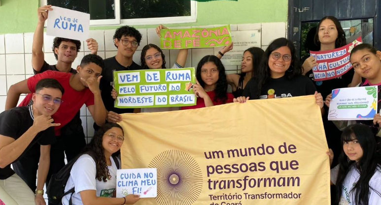 A imagem mostra um grupo de jovens em uma instituição educacional, segurando cartazes e uma faixa com mensagens de apoio a causas ambientais e sociais. Entre os dizeres, estão frases como "Vem de Ruma pelo futuro do Nordeste do Brasil", "Amazonia depende", "Natureza não é cultura, é ensino no Nordeste", "Cuida no clima, meu fi!" No centro está a bandeira do Território Transformador do Ceará.