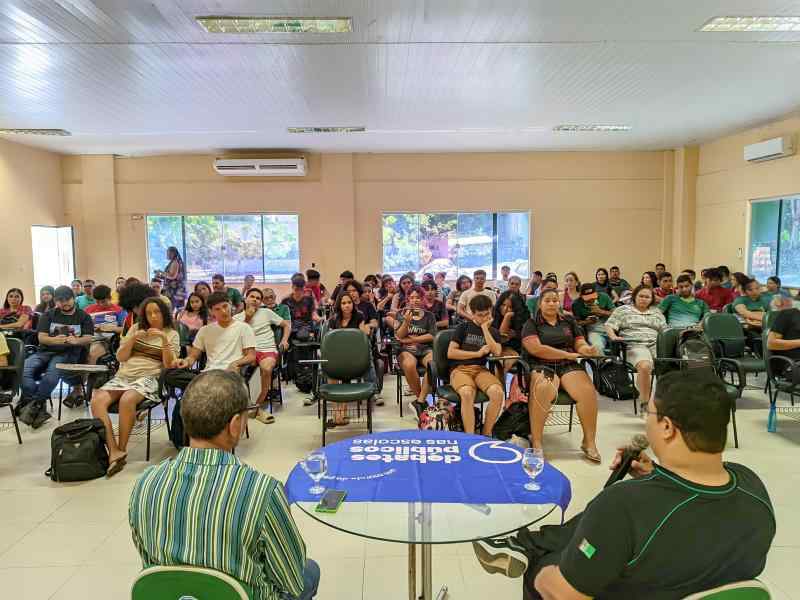 A imagem mostra uma sala de conferências onde um grupo grande de pessoas está reunido. O público, composto principalmente por jovens, está sentado em cadeiras distribuídas em fileiras. Na frente da sala, dois homens estão sentados de costas para a câmera. Um deles, segurando um microfone. Há uma mesa redonda de vidro com a bandeira dos Debates Públicos nas escolas.  A sala possui amplas janelas ao fundo, permitindo a entrada de luz natural e uma vista das árvores externas.