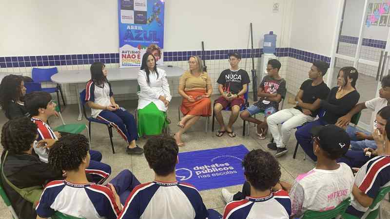 Sala com pessoas sentadas em circulo e uma bandeira estendida no meio, com o simbolo dos Debates Publicos nas Escolas