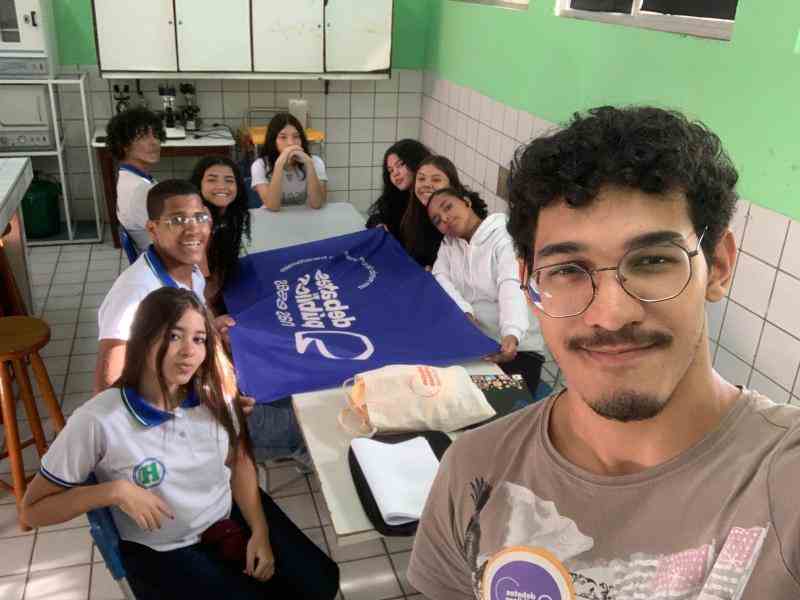 Grupo de jovens reunidos em um sala, em  volta de uma mesa,com a bandeira dos Debates Publicos nas Escolas, foto em formato Selfie.