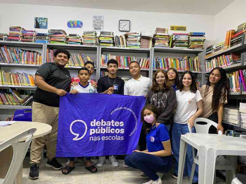Grupo de jovens reunidos numa biblioteca, posando para foto com uma bandeira dos debates publicos nas Escolas
