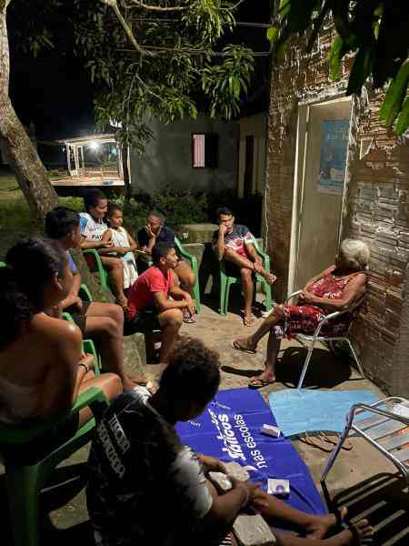  Foto de uma roda de conversa à noite, em uma área externa, com uma senhora idosa de cabelo branco, sentada em uma cadeira, compartilhando histórias com crianças e adolescentes, que estão atentos, sentados em cadeiras verdes. No chão, uma canga azul do projeto Debates Públicos nas Escolas