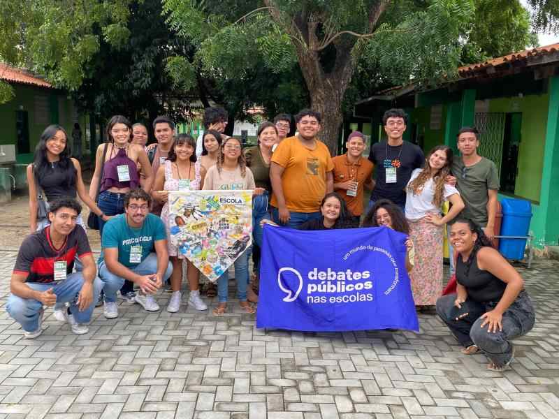 Grupo de jovens reunidos com  uma bandeira dos Debates Publicos Nas Escolas estendida sob o chão.