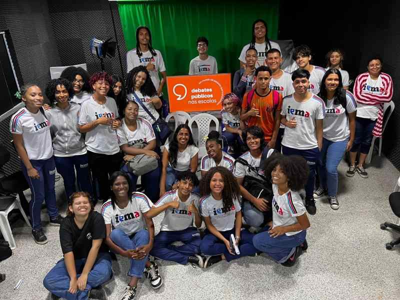 Grupo de jovens reunidos em um sala, uma bandeira dos Debates Publicos Nas Escolas estendida sob o chão.