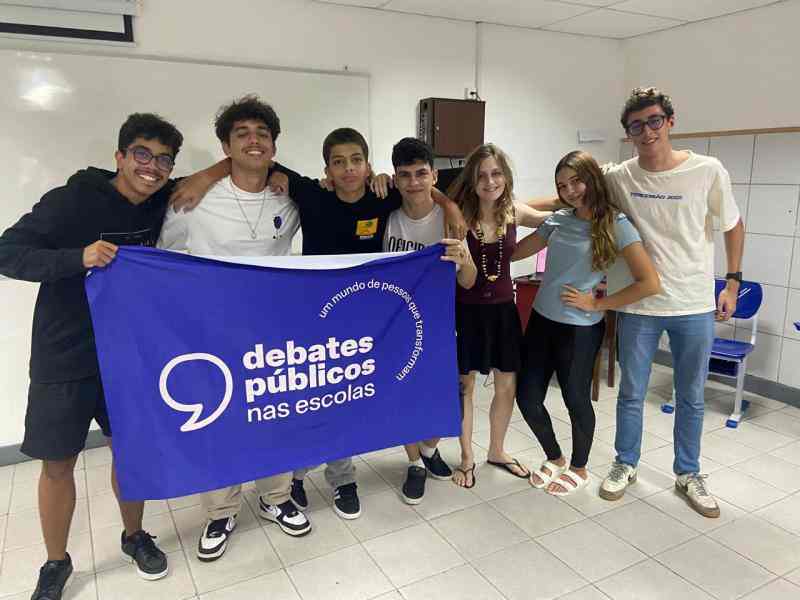 Grupo de jovens reunidos em um sala, uma bandeira dos Debates Publicos Nas Escolas estendida sob o chão.