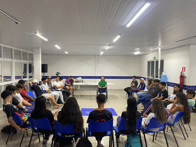 Grupo de jovens sentados em cadeiras formando um semicírculo em uma sala de aula. Ao centro, há a bandeira dos Debates Públicos