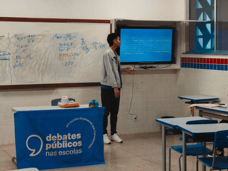 Jovem em uma sala de aula ao lado de um quadro branco, uma mesa com a bandeira dos Debates Públicos e uma TV. 