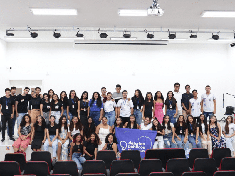 Grupo de jovens reunidos em um auditório. Ao centro, alguns deles seguram a bandeira dos Debates Públicos