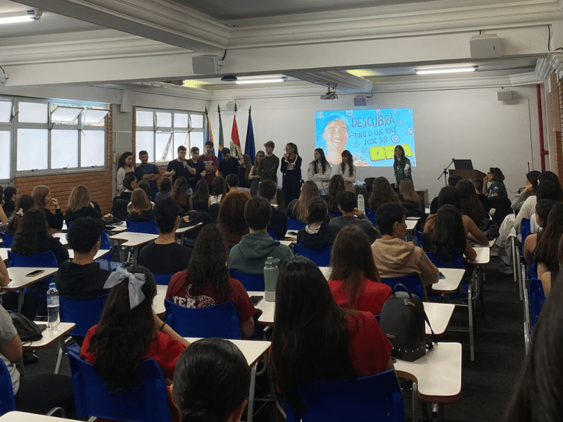 Sala de aula com estudantes sentados em carteiras, enquanto um grupo está à frente conduzindo uma atividade.