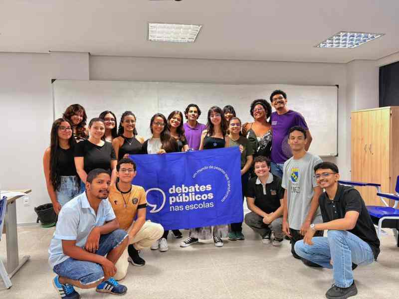 Grupo de jovens reunidos em uma sala de aula posando para a foto. Ao centro, elas seguram a bandeira roxa dos Debates Públicos nas Escolas